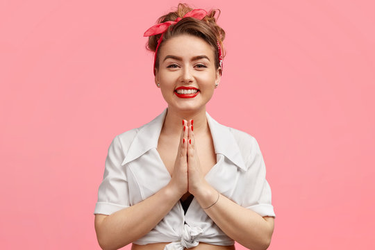 Photo of glad female with cheerful expression, keeps palms pressed together, asks for help, smiles broadly, has white teeth and red lips, isolated over pink background. People and praying concept