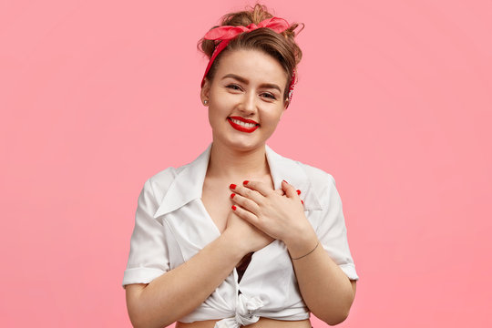 Attractive young female model with red lips, being touched by something, keeps palms on heart, expresses gratitude, wears white blouse, isolated over pink background, has make up. Emotions concept