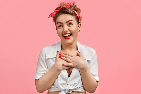 Oh, I am so touched! Lovely pleased excited female has joyful expression, keeps hands on heart, wears white top, glad to recieve pleasant words, keeps both palms on chest isolated over pink background