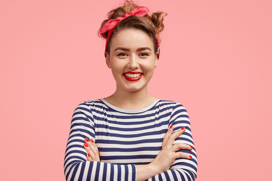 Happy female with positive expression, keeps hands crossed, dressed in striped jacket and stylish headband, poses against pink background, glad to see her photo on poster. People and emotions