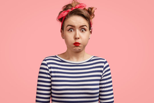 Indoor shot of beautiful European female in retro style clothes, blows cheeks, stares with popped eyes at camera, being amazed by shocking news, isolated over pink background. Facial expressions