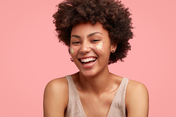 Funny happy female with Afro hairstyle, has broad smile, shows white perfect teeth, dressed in casual outfit, has sparkles on cheeks, isolated on pink background. Positive young African American woman