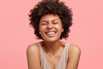 Happpiness concept. Overjoyed pleased dark skinned young female with Afro curly hairstyle, has broad smile, keeps eyes closed from pleasure, enjoys something great, stands against pink wall. Glitter