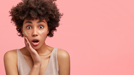 Horizontal shot of surprised young African American female with jaw dropped and widely opened eyes, has crisp dark hair, isolated over pink background with copy space for your advertisement.
