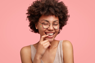 Pleased adorable African American female smiles gently at camera, keeps fore finger near teeth, closes eyes, dreams and imagines something pleasant, isolated over pink background. Emotions concept