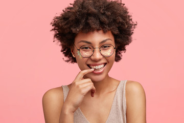 Portrait of happy African American female with joyful expression, broad smile and white teeth, has bright sparkles on face, stands against pink background, being in great mood after outdoor walk