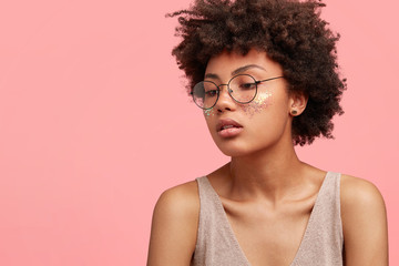 Sideways shot of beautiful thoughtful female concentrated aside, wears round glasses, has glitter on cheeks, Afro hairstyle and dark skin, dressed in oversized casual t shirt, stands against pink wall