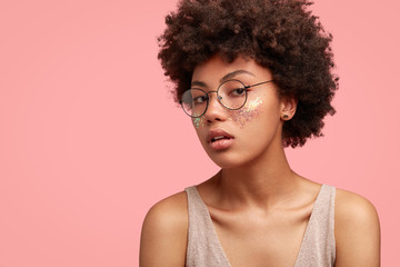 Profile of charming African American woman with serious expression, looks seriously through spectacles, has glitter on face, pure healthy skin, stands against pink wall with copy space for your text