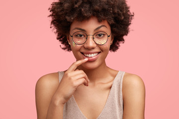 Adorable satisfied dark skinned female youngster has crisp hair, raises eyebrows, smiles gently, listens pleasant story from friend, isolated over pink background. Ethnicity and emotions concept