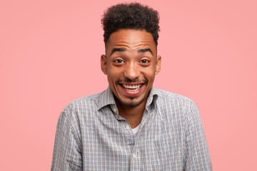 Cheerful dark skinned guy has trendy haircut, giggles happily, dressed in checkered shirt, stands against pink background, models in studio. Smiling African American man expresses positive emotions