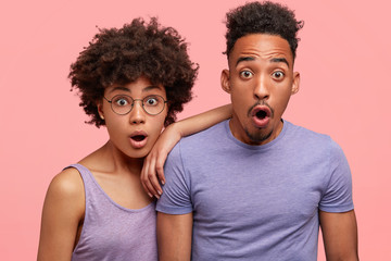 Indoor shot of handsome male and his girlfriend look with terrified expressions, keep jaws dropped, react on awful news, express surprisement, isolated over pink background. Dark skinned partners