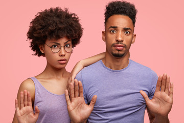 Horizontal shot of serious African American wife and husband show stop gesture, deny something with nagative facial expression, stand against pink background. Companionship and body language