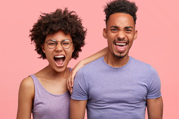 Furious dark skinned female and male shout angrily, have quarrel with neighbours who are very noisy, complain about something, keep mouthes wide opened, isolated on pink background. Negative emotions
