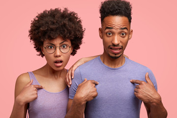 Why should we do this? Embarrassed dark skinned African American female and her male friend, point at themselves, wonder to be chosen for making project by professor, isolated on pink background
