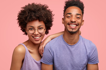 Cute cheerful dark skinned female and male couple smile peasantly at camera, have joyful looks, stand close to each other, pose against pink wall, express happiness. True friendship concept.