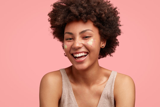 Funny happy female with Afro hairstyle, has broad smile, shows white perfect teeth, dressed in casual outfit, has sparkles on cheeks, isolated on pink background. Positive young African American woman