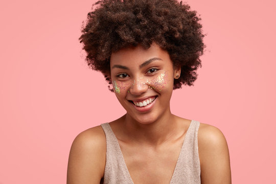 Joyful young African American female teenager has broad smile, shows white teeth, looks happily at camera, has bright sparkles on her face, isolated over pink background. Positiveness concept