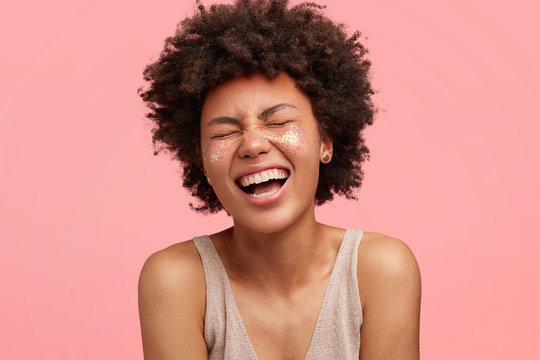 Close up portrait of happy African female laughs joyfully, closes eyes and smiles broadly, has glitter on cheeks, dressed in oversized t shirt, poses against pink background, expresses positiveness