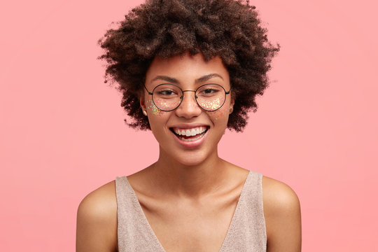 Headshot of pretty African American female has face decorated with glitter, smiles pleasantly at camera, wears round spectacles, isolated over pink background. People, beauty and happiness concept