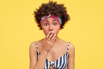 Naklejka premium Young attractive African American female with scared expression, covers mouth and looks in puzzlement, can`t believe in latest awful news about friend, wears headband and striped casual t shirt