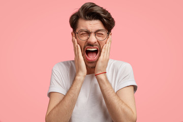 Stressful desperate European male keeps mouth widely opened and eyes shut, shouts angrily, expresses negative emotions, scratches cheeks, isolated over pink background. People and reaction concept