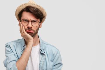 Indoor shot of unhappy unshaven male with stubble, has gloomy expression, holds chin, wears straw hat and denim shirt, poses against white background with blank copy space for your advertisement