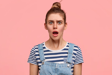 Emotive frustrated surprised European female teenager looks with puzzlement at camera, dressed in casual t shirt and denim overalls, poses against pink wall, receives bad news from interlocutor