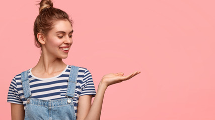 Lovely happy female teenager in denim dungarees, has hair combed in knot, pretends holding something, raises palm against pink wall with blank space for your promotional text or advertisement