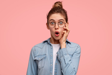 Portrait of amazed cute female stares at camera, keeps mouth wide opened, can`t believe in her failure, stands against pink background, dressed in denim jacket. People, reaction and emotions concept