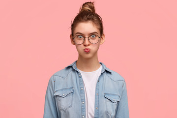 Indoor shot of good looking female blows cheeks, has funny expression, wears round spectacles, dressed in casual denim jacket, gasps from relief, isolated over pink background. Facial expressions