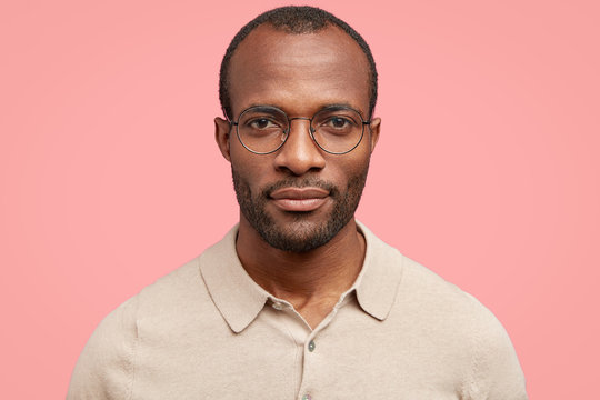 Close up portrait of handsome self assured dark skinned male, looks directly at camera, wears round spectacles, isolated over pink background. Headshot of intelligent student ready to present project