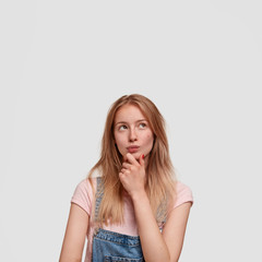 Charming Caucasian female with light hair, picks something in mind, thinks as needs to make difficult choice, dressed in fashionable denim overalls, isolated over white blank wall, holds chin