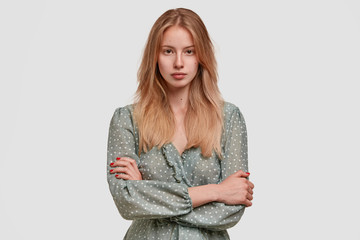 Waist up portrait of pretty light haired confident female keeps hands crossed, wears polka dot blouse, has appealing appearance, isolated over white concrete wall. People, beauty and style concept