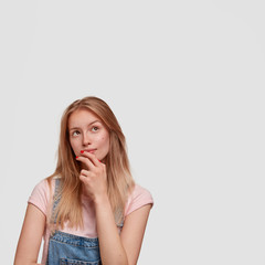 Vertical shot of attractive female has thoughtful focused expression upwards, concentrated on something, holds chin, dressed in denim dungarees, poses against white blank wall. What should I choose?