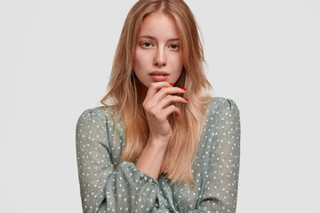 Cropped shot of lovely young European female with red manicure, wears stylish blouse, holds chin, looks thoughtfully at camera, poses against white wall, thinks about participating in fashion show