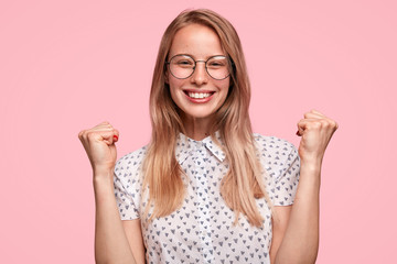 Happy beautiful student in eyewear rejoices success and achieving great results in studying, clenches fists, has broad charming smile, isolated over pink background. Yes, I did it! Triumph concept