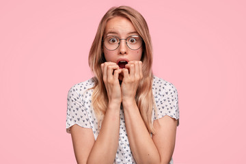Embarrassed European young female stares at camera and bites finger nails with worried expression, doesn`t believe in her failure, wears spectacles and t shirt, stands against pink background