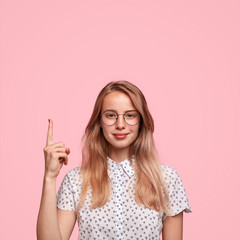 Pleased lovely young female with light hair, healthy skin, wears spectacles, indicates with fore finger upstairs, shows blank space, wears elegant blouse, being impressed by something amazing
