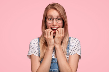 Surprised beautiful young female has embarrassed expression, looks nervously at camera, bites finger nails, hears unexpected news, dressed in denim dungarees, stands against pink background.