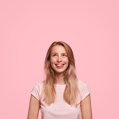 Happy young woman with appealing appearance looks upwards with charming smile, sees something pleasant, dressed in casual t shirt, isolated over pink wall. Monochrome, people, happiness concept