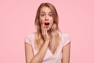 Stunned beautiful female model expresses surprisement as recieves intriguing unexcpected information, keeps jaw dropped, wears casual clothes, poses against pink blank wall. People and stress