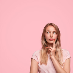 Vertical shot of thoughtful European female looks upwards, keeps fingers on cheek, contemplates about future wedding with boyfriend, dressed casually, has weekend, isolated on pink wall. Monochrome