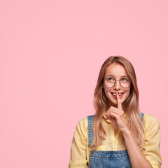 Positive female youngster dressed in overalls, has cheerful expression while makes silence gesture, looks mysteriously upwards, asks to keep inforamtion in secret, isolated over pink background