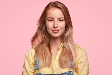 Lovely satisfied female student happy to finish university and makes plans for future, dressed in yellow shirt and dungarees, has pleased expression, looks directly at camera, isolated on pink wall