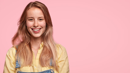 Studio shot of happy Caucasian female has gentle smile, rejoices successful day, has long hair, poses over pink blank wall with free space for your advertising content. People, beauty, emotions