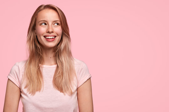 Horizontal shot of pretty happy female with broad smile, being in high spirit, dreams or imagines something pleasant, stands against pink wall with copy space. People, positive emotions concept