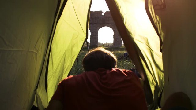 Young man backpacker exits camping tent in front of roman aqueduct arches in parco degli acquedotti park ruins in rome at sunset sticks in ground shot from inside tent camera slow motion pov