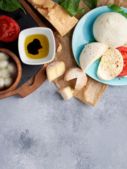 Italian cheeses. Mozzarella Buffalo, crottin, balls, Stracciatella, basil leaves, tomatoes, olive oil with vinegar on gray background. Top view, copy space.
