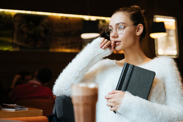 young beautiful freelancer girl in glasses and a white jacket is sitting in a cafe, holding her...