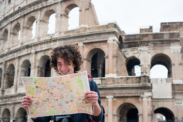 Handsome young traveler looking at tourist map in Rome in front of Colosseum. Backpacker with camera and tourist map